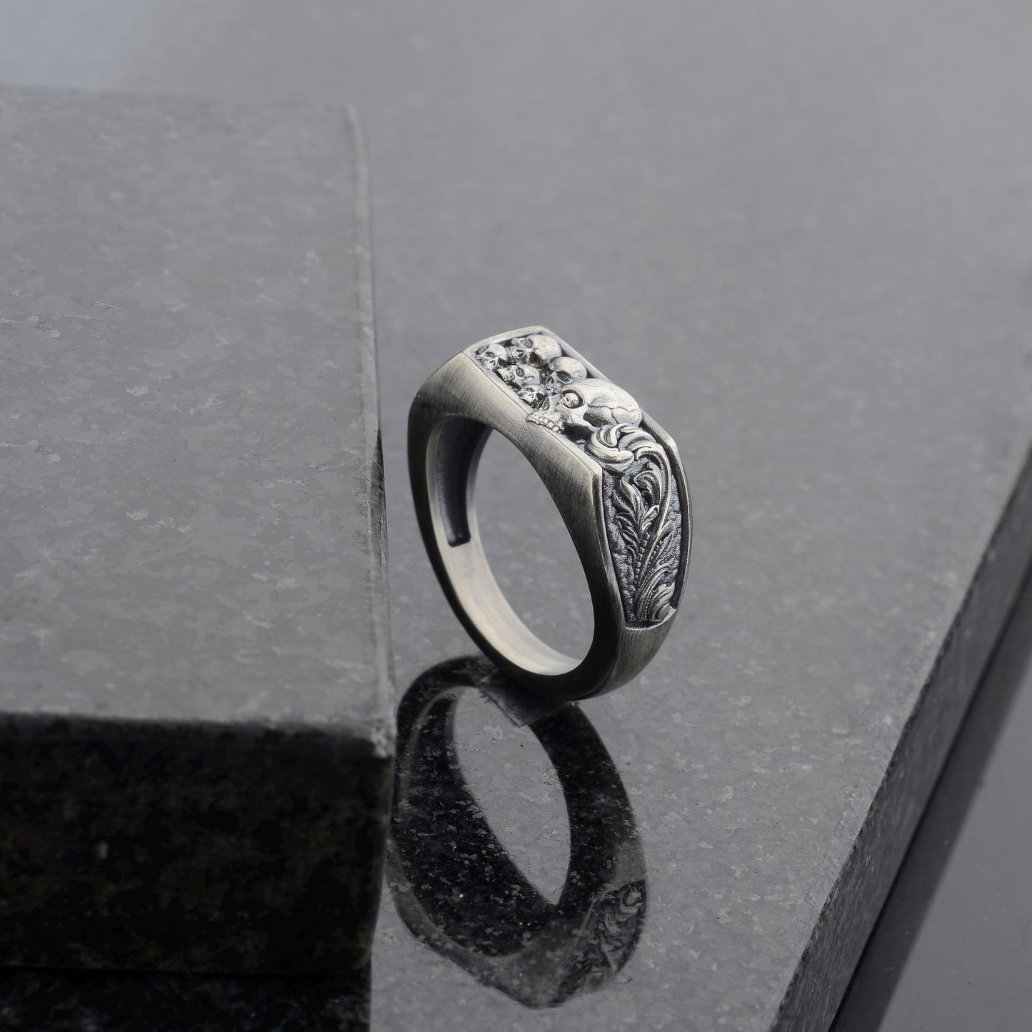 May include: A silver-toned ring with a rectangular face featuring a detailed relief design. The design includes skulls and ornate floral elements. The ring is set against a dark, reflective surface.