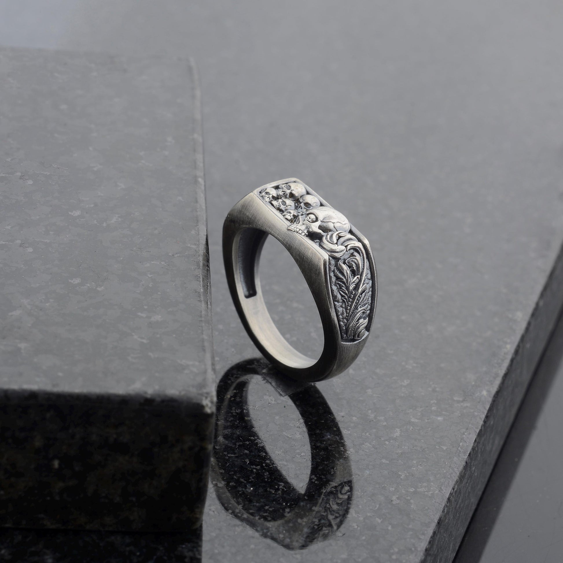 May include: A silver-toned ring with a rectangular face featuring a detailed relief design. The design includes skulls and ornate floral elements. The ring is set against a dark, reflective surface.