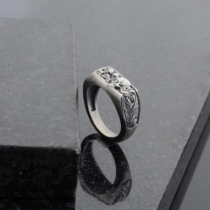 May include: A silver-toned ring with a rectangular face featuring a detailed relief design. The design includes skulls and ornate floral elements. The ring is set against a dark, reflective surface.