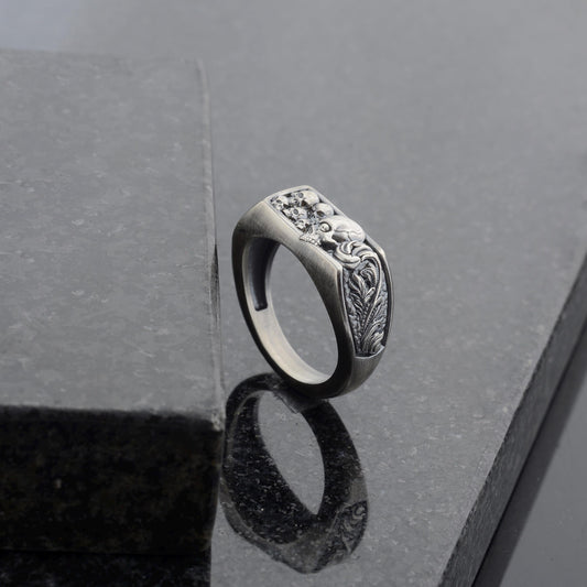 May include: A silver-toned ring with a rectangular face featuring a detailed relief design. The design includes skulls and ornate floral elements. The ring is set against a dark, reflective surface.