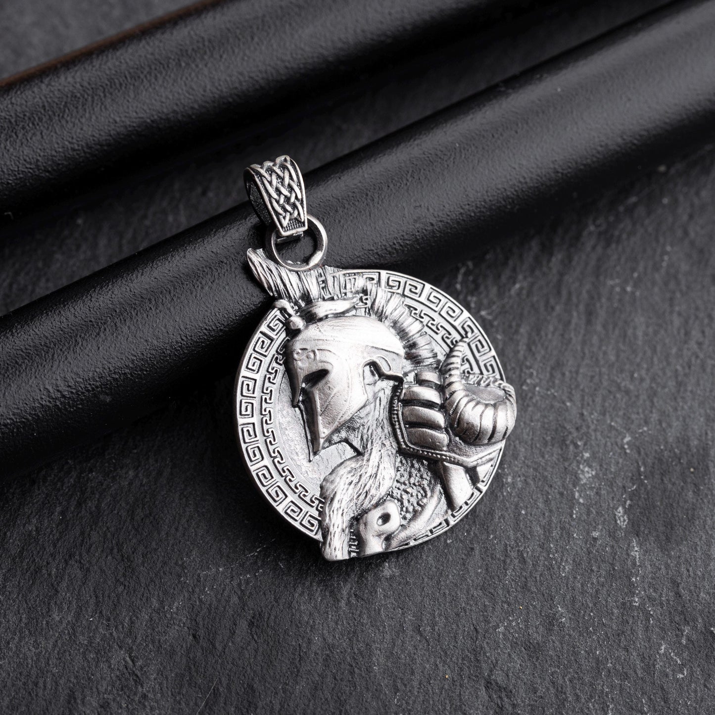 Gladiator Pendant in silver featuring a detailed helmeted warrior profile with a ram's head and intricate braided bail, set against a patterned circular background.