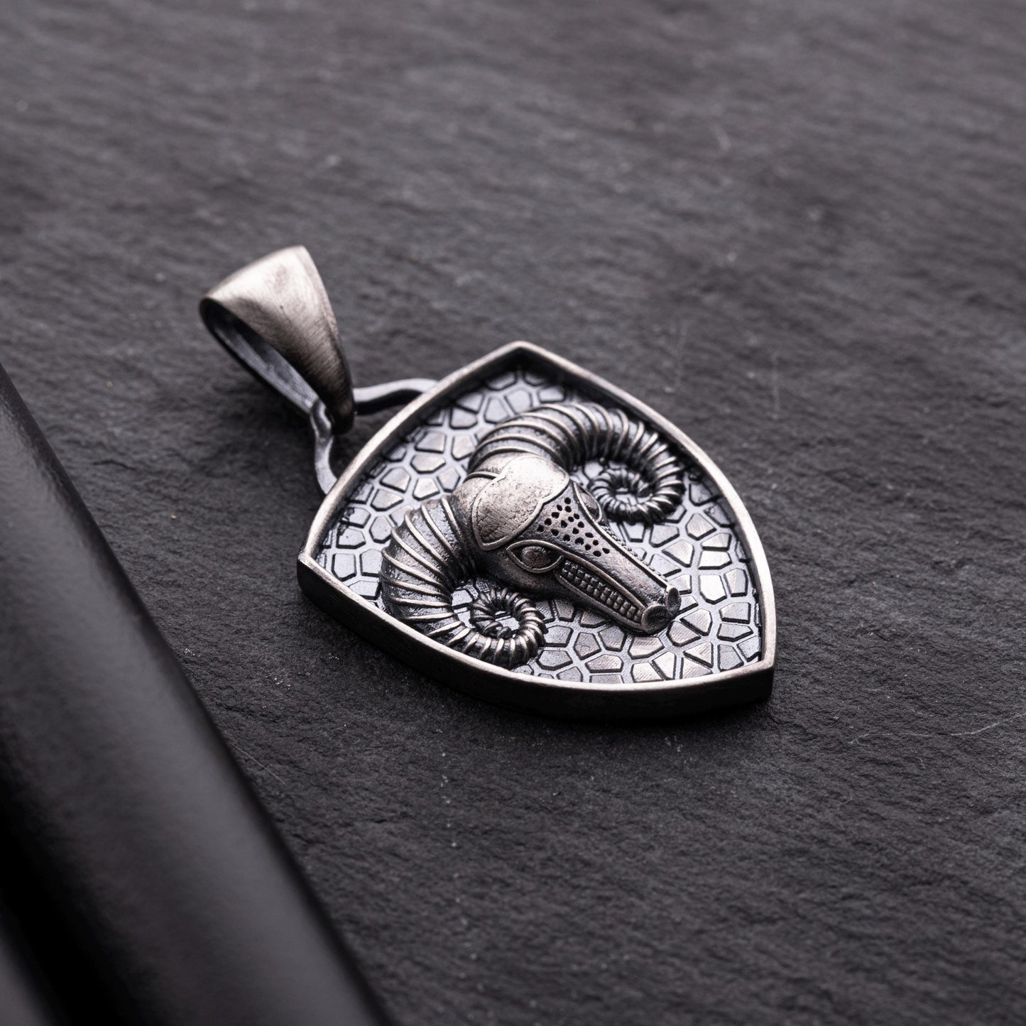Aries Ram Pendant featuring a detailed ram's head with curled horns set on a textured silver shield-shaped background, showcasing intricate craftsmanship on a dark surface.