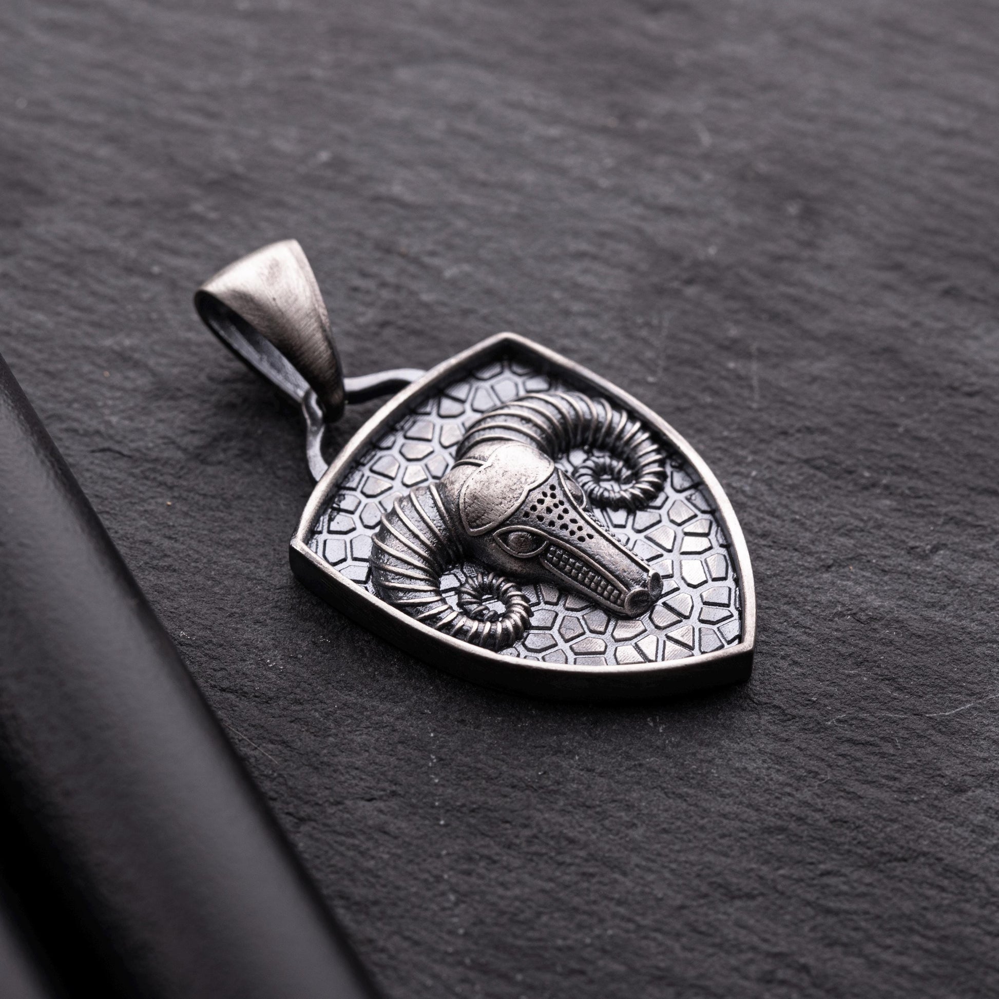 Aries Ram Pendant featuring a detailed ram's head with curled horns set on a textured silver shield-shaped background, showcasing intricate craftsmanship on a dark surface.