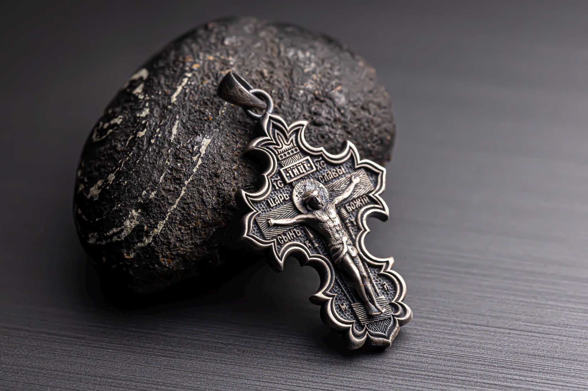 Jesus Crucifix Pendant in detailed silver design resting against a textured dark stone on a smooth black surface, showcasing intricate craftsmanship.