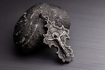 Jesus Crucifix Pendant in detailed silver design resting against a textured dark stone on a smooth black surface, showcasing intricate craftsmanship.