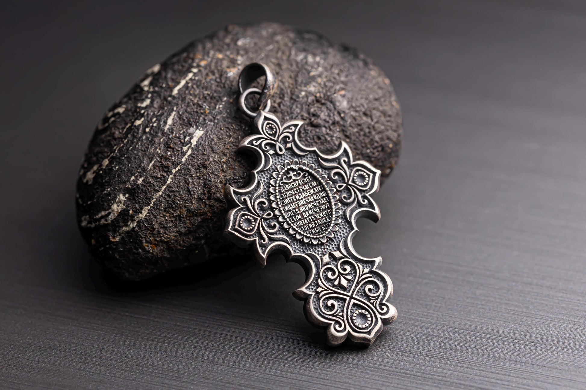 Intricately designed Jesus Crucifix Pendant in silver tone resting against a textured dark stone, featuring detailed patterns and delicate scrollwork for a stylish look.