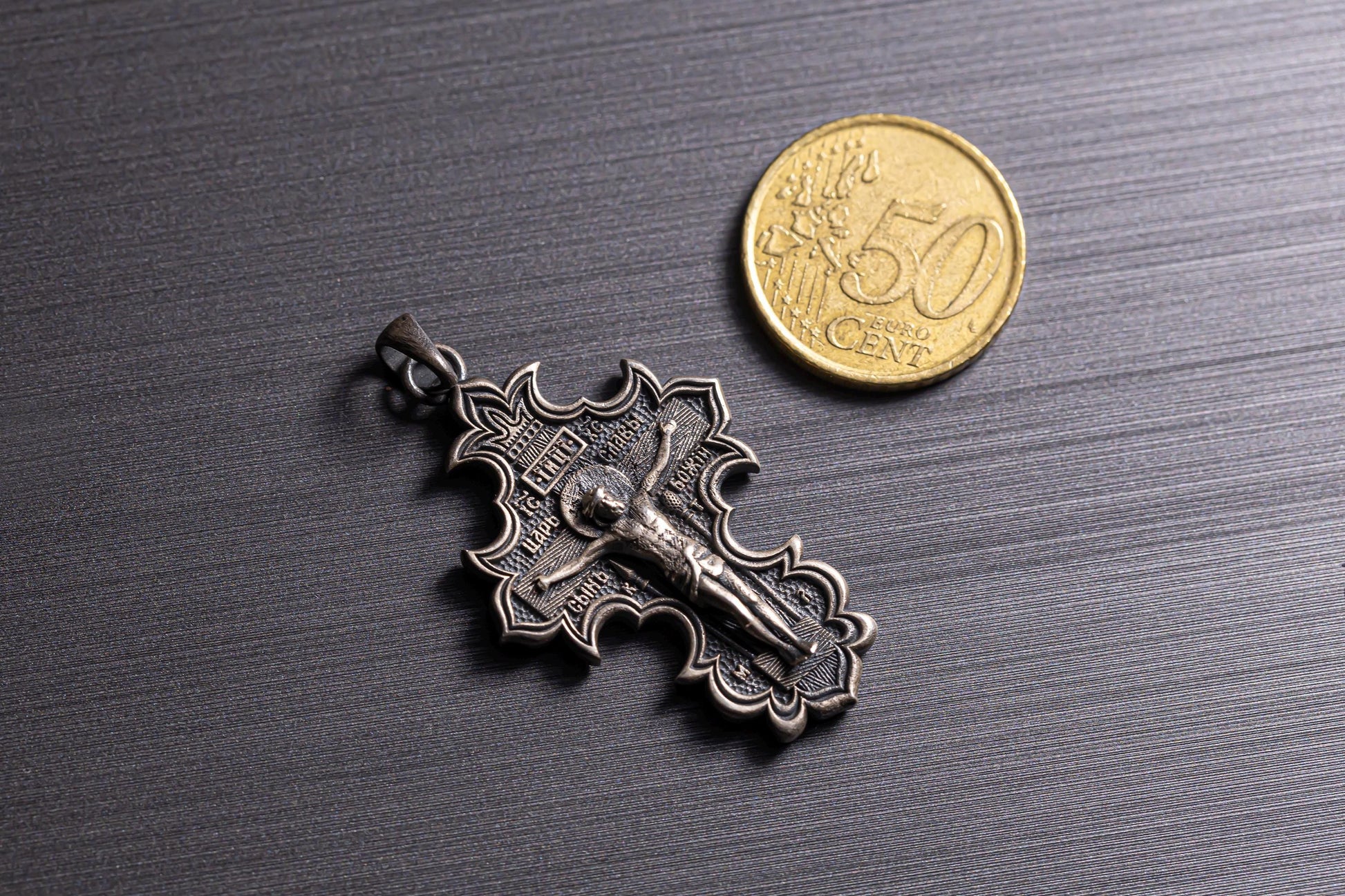 Jesus Crucifix Pendant in detailed silver metal with ornate edges, shown next to a 50 cent coin for size comparison on a dark textured surface.