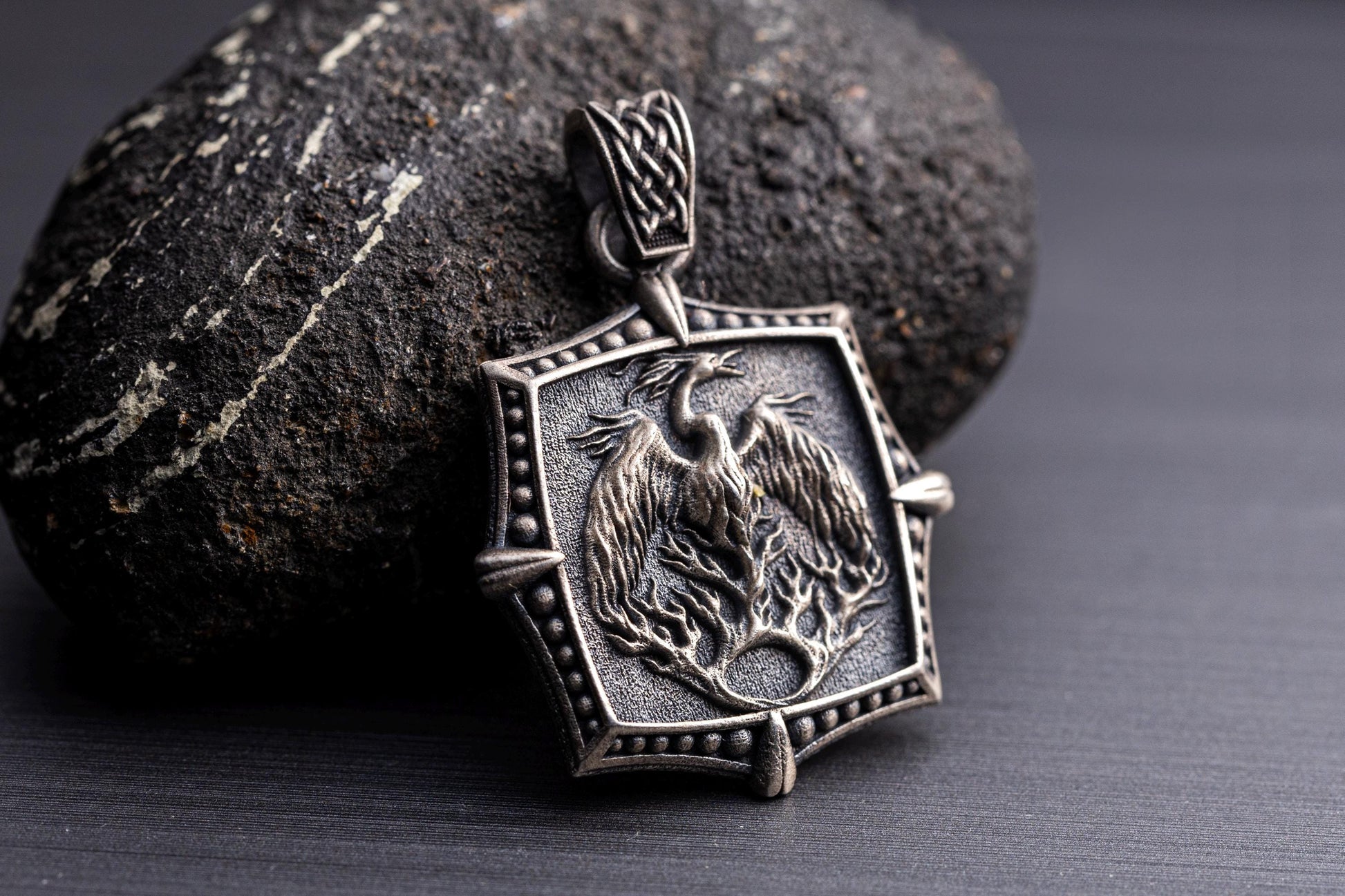 Silver Phoenix Pendant featuring an intricately detailed phoenix rising from flames on a textured silver background, displayed against a dark stone surface.