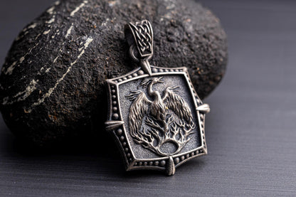 Silver Phoenix Pendant featuring an intricately detailed phoenix rising from flames on a textured silver background, displayed against a dark stone surface.