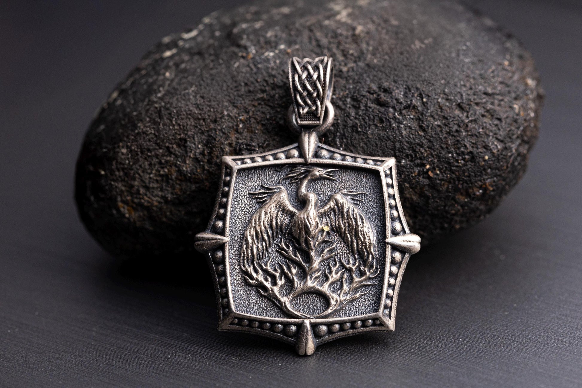 Silver Phoenix Pendant featuring detailed phoenix rising from flames design in oxidized silver finish, resting on a dark textured stone background.