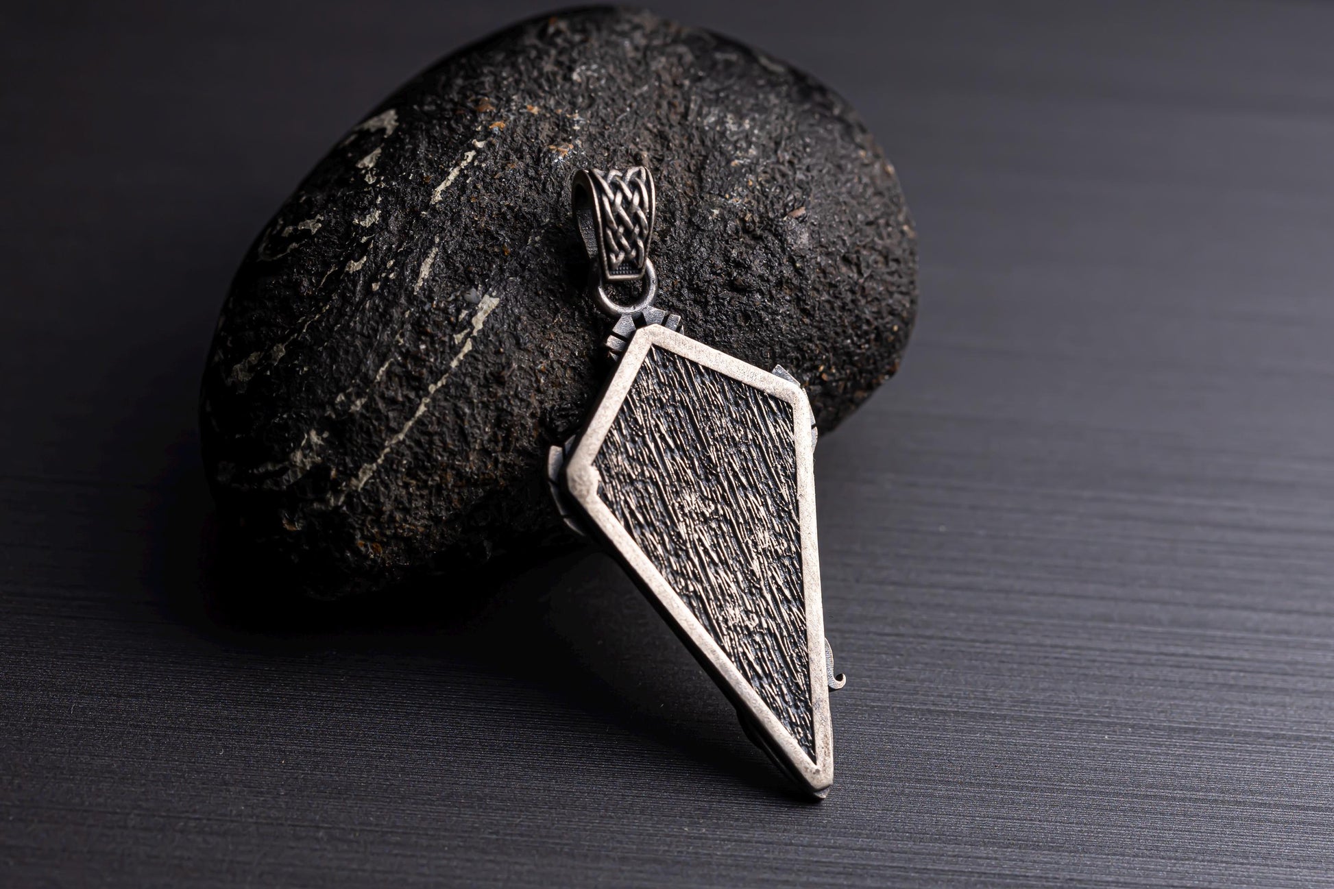 Sterling Silver Dragon Arrowhead Pendant resting on a textured black stone, featuring a detailed, rugged design with a silver frame and intricate chain necklace loop.