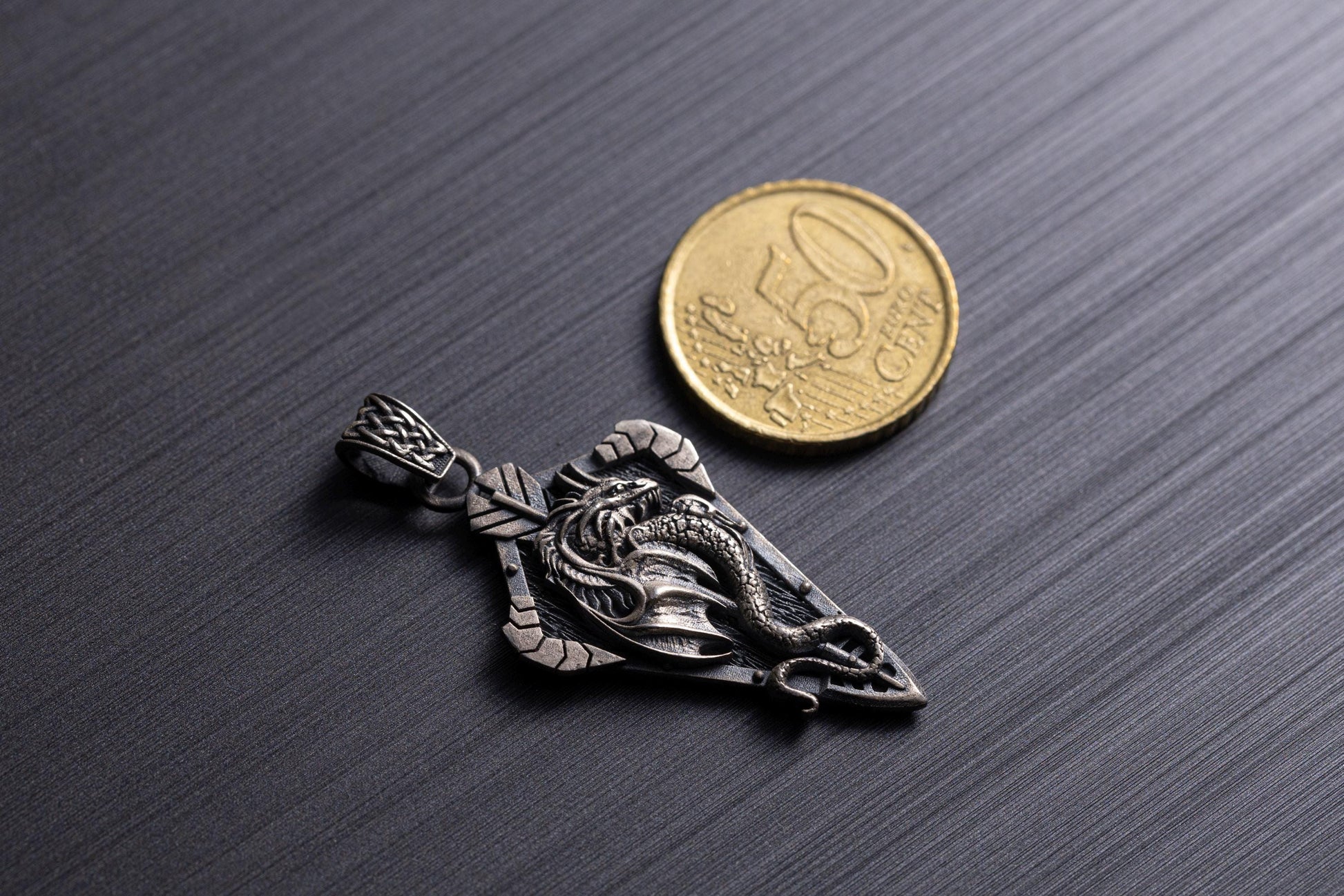 Sterling Silver Dragon Arrowhead Pendant with detailed dragon design displayed next to a 50 cent coin for size reference on a dark textured surface.